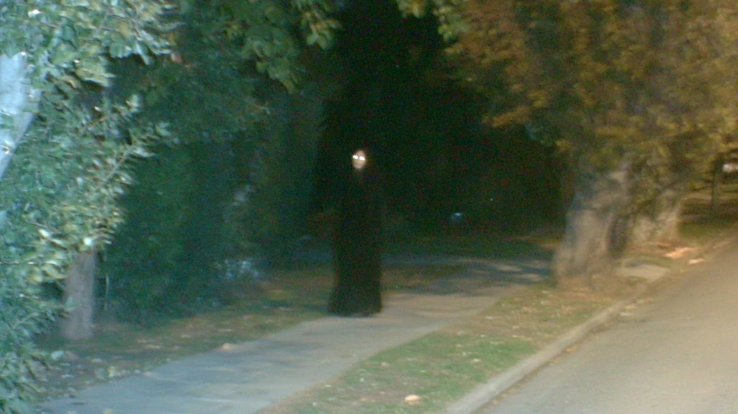 Uma entidade feminina espectral vestindo uma longa roupa preta está de pé em uma calçada suburbana cercada por árvores à noite