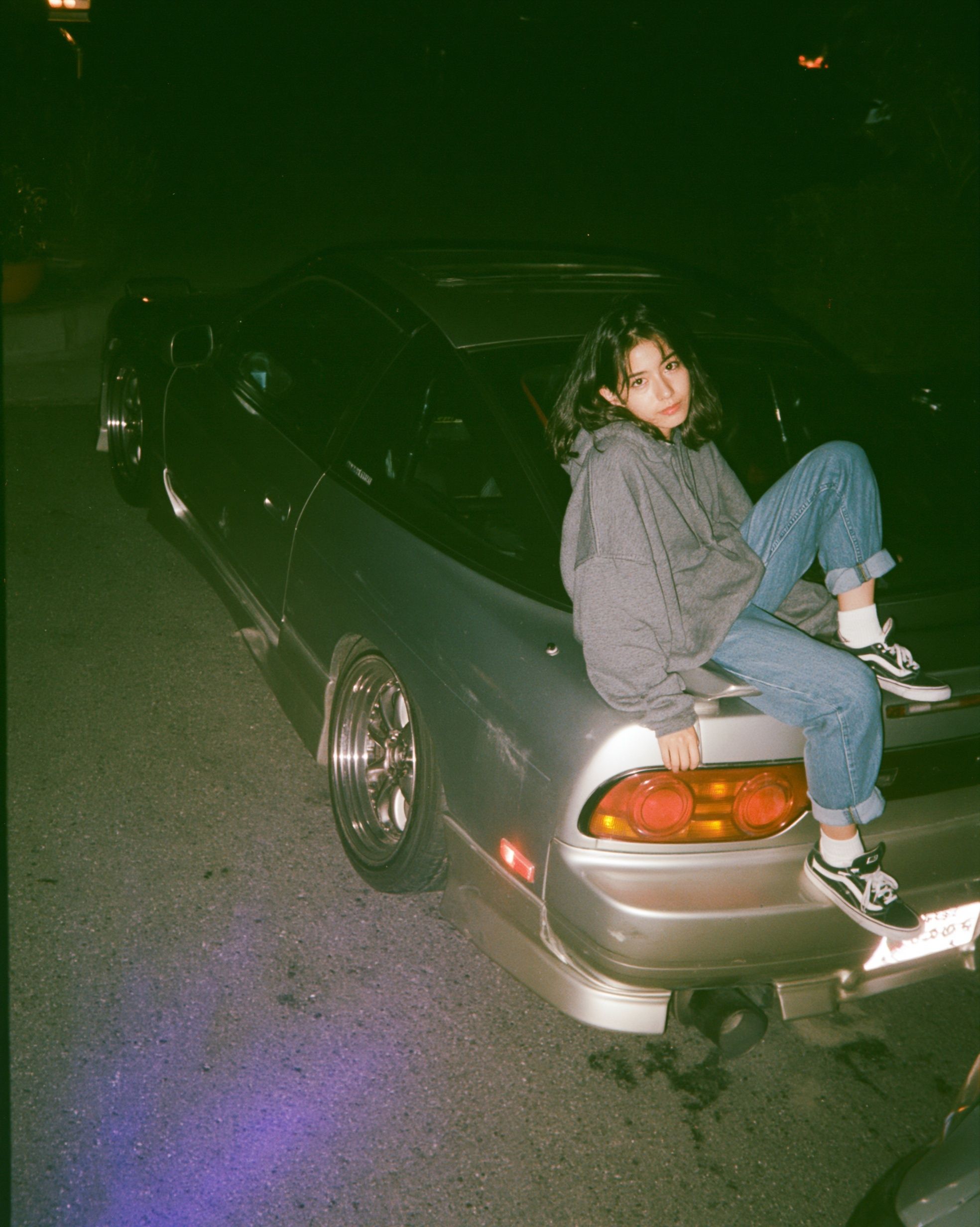 A nostalgic 1990s Japanese street car night scene with a young woman sitting on the rear of a Nissan 180SX