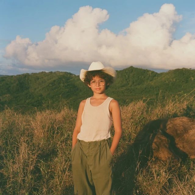 A young boy wears a white cowboy hat and sleeveless white tank top standing in a grassy field