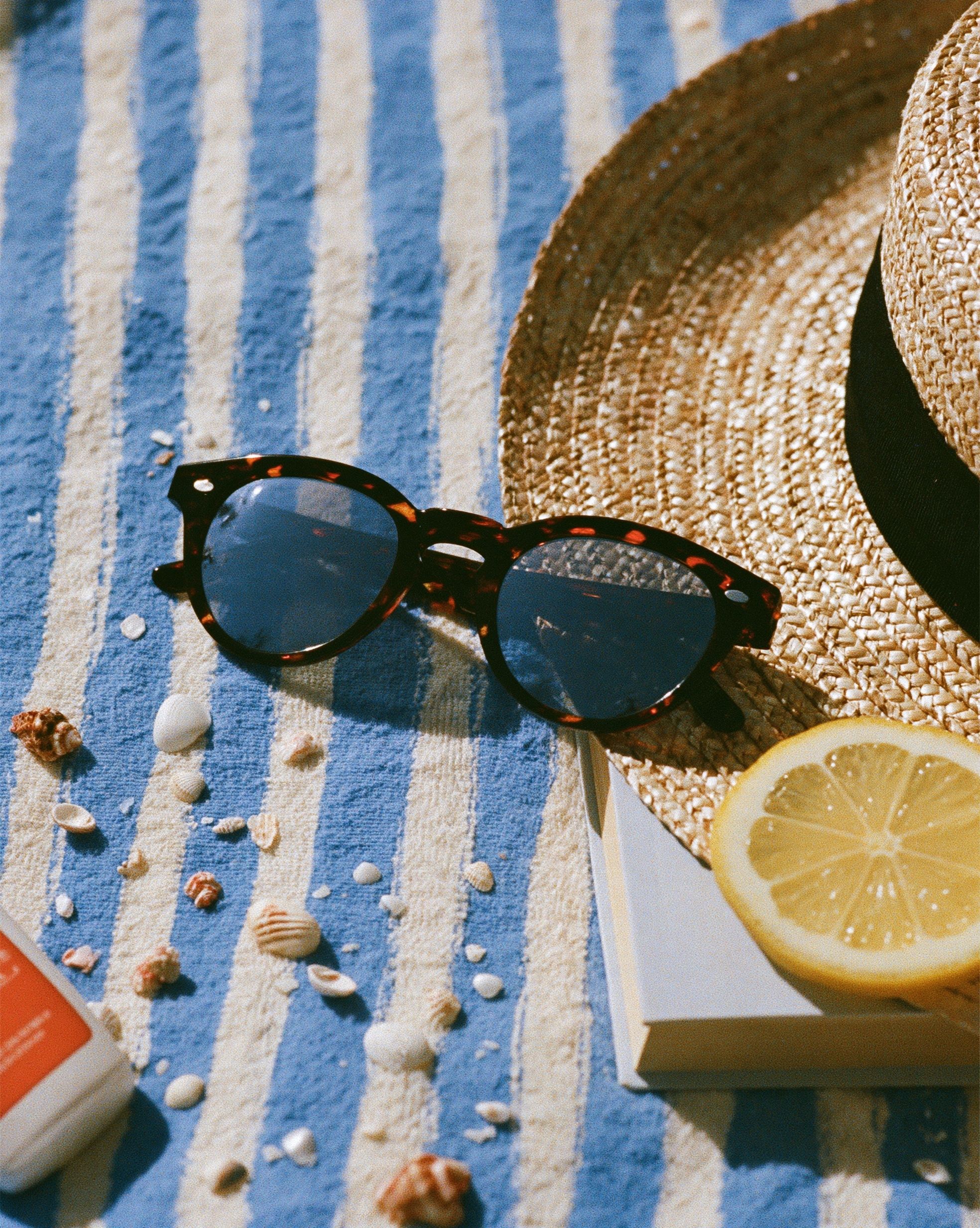 Flachliegende Sommerstrand-Essentials einschließlich Sonnenbrille, Sonnencreme, Strohhut, Taschenbuch, Zitronenscheibe, Muscheln und gestreifter Handtuch